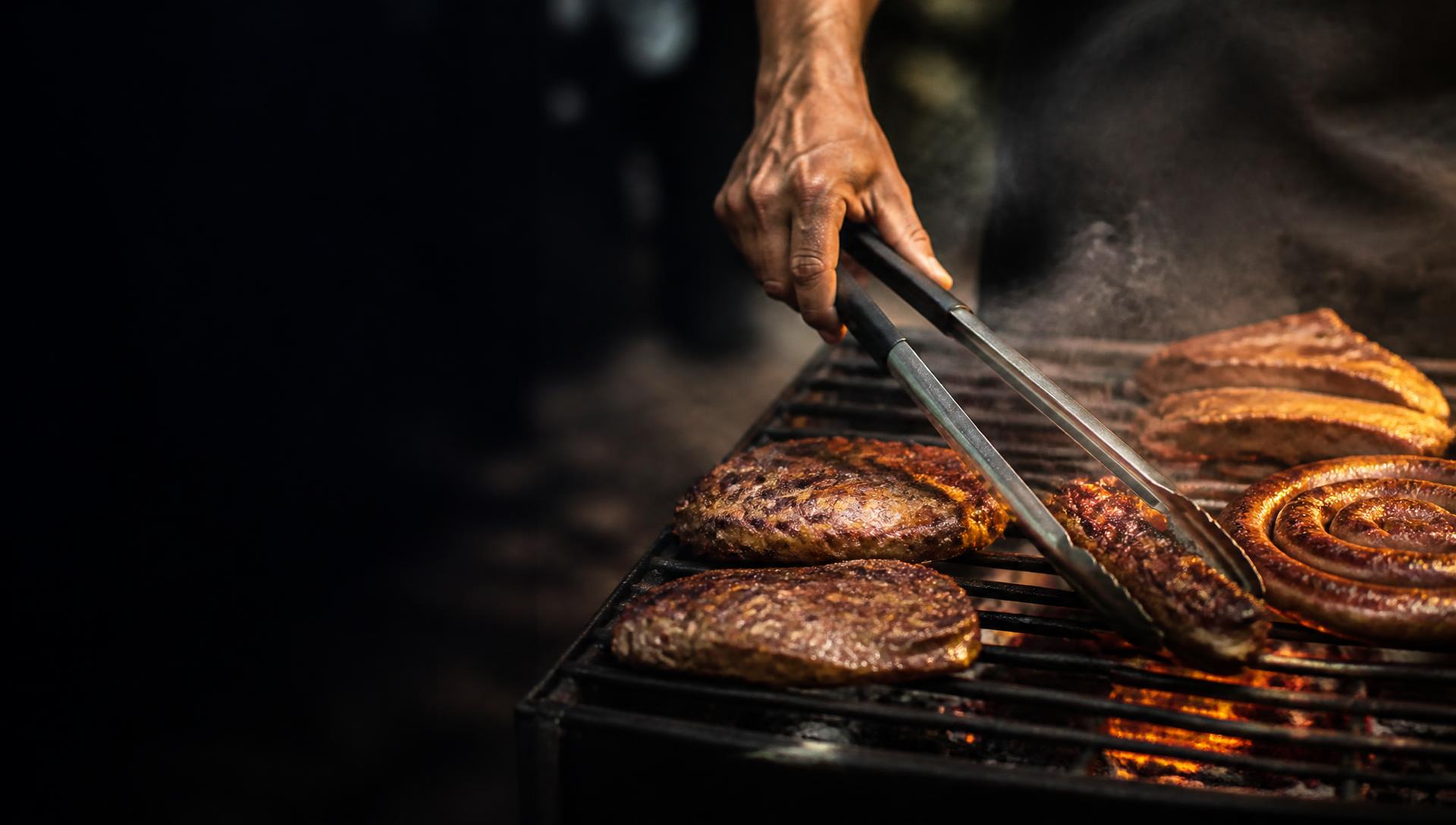 Braaier turning steak and boerewors over the coals with a pair of long braai tongs